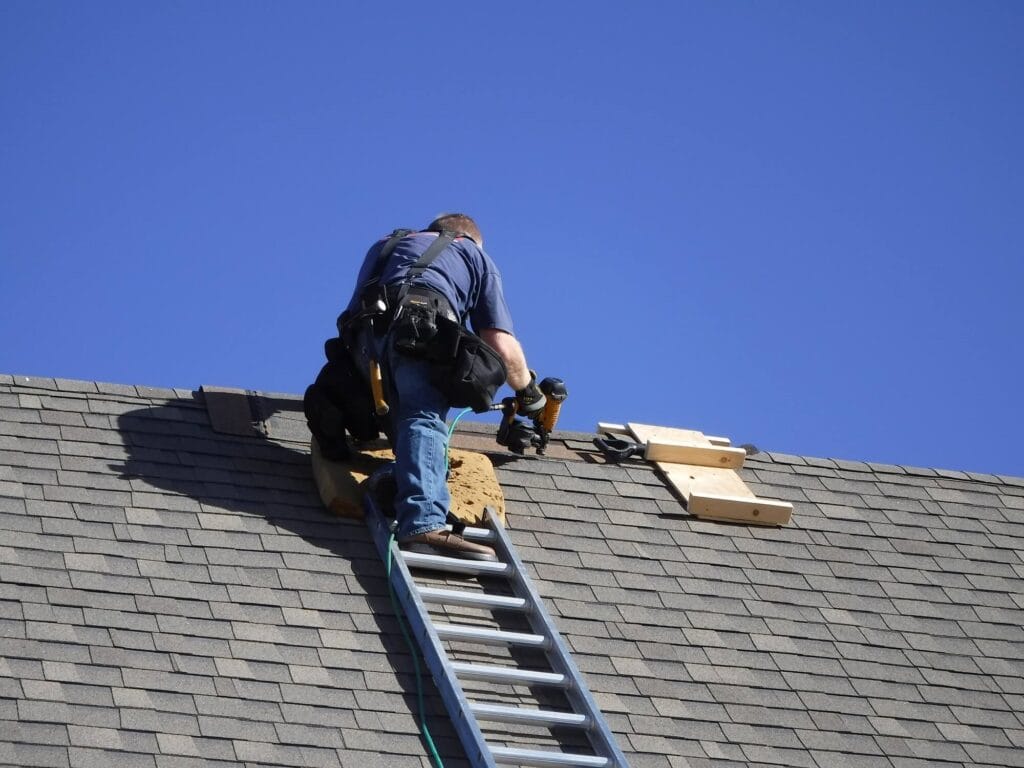 Roofing worker installing or repairing shingles on a sloped roof under a clear blue sky.