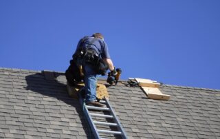 Roofing worker installing or repairing shingles on a sloped roof under a clear blue sky.