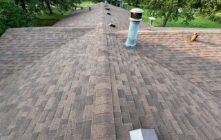 View of a brown shingle roof with vents and a chimney, surrounded by green trees.